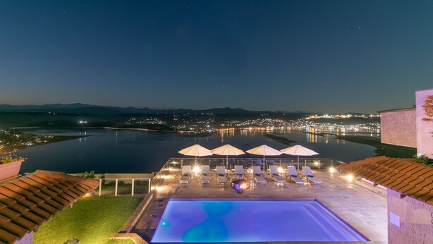 A rooftop infinity pool with lounge chairs and umbrellas, overlooking a cityscape and water body at night. - Villa Castollini (Knysna)