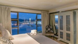 A hotel room with a large bed, a chair, a cabinet, and a view of a cityscape at night. - Villa Castollini (Knysna)