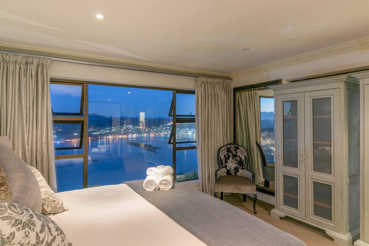 A hotel room with a large bed, a chair, a cabinet, and a view of a cityscape at night.
