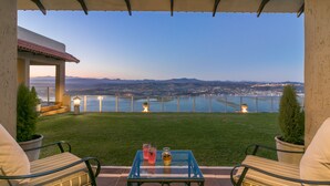 A patio with a table, two chairs, and a view of a body of water and mountains. - Villa Castollini (Knysna)