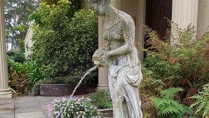 A stone statue of a woman pouring water, surrounded by a circular stone basin, set in a garden with various plants and a building in the background. - Villa Castollini (Knysna)