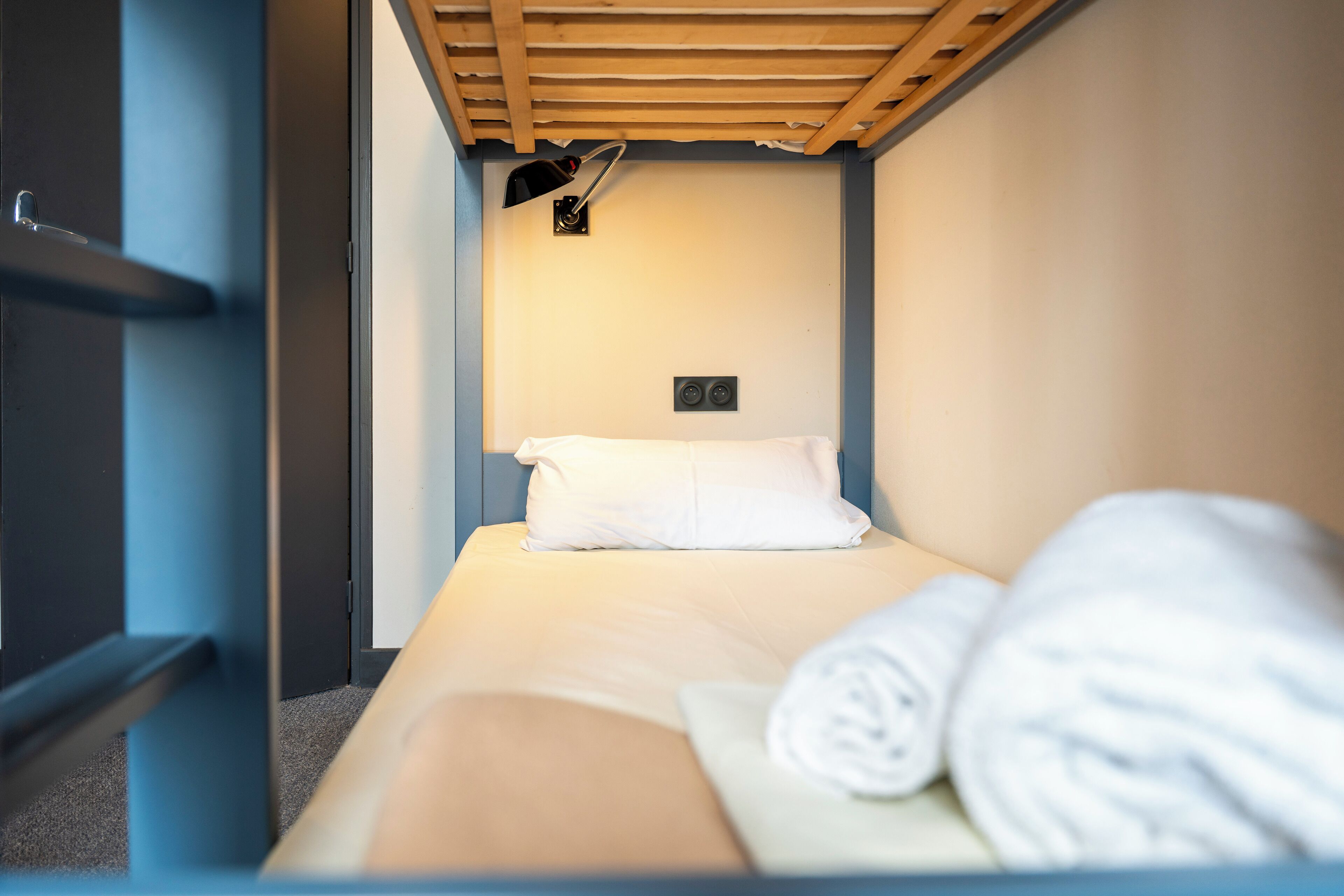 A single bed with a white pillow and a rolled towel, a small wall-mounted light, and a power outlet on the wall.