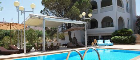 A swimming pool with lounge chairs and umbrellas, a covered seating area, and a building with multiple balconies.