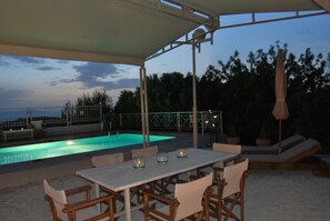 Uma área de piscina externa com um terraço coberto, assentos e vista para o mar.