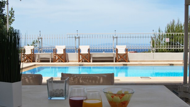 Une table placée au bord de la piscine, avec des boissons et un saladier de fruits, donnant sur une piscine entourée de chaises et d’une clôture.