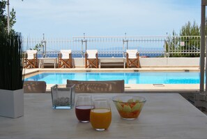 Mesa junto a la piscina con bebidas y frutero, con vista a una piscina con sillas y una valla.