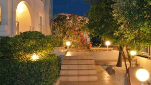 Un espace extérieur bien éclairé, avec des marches menant à l’entrée d’un bâtiment, entouré de verdure et illuminé par des lampadaires.