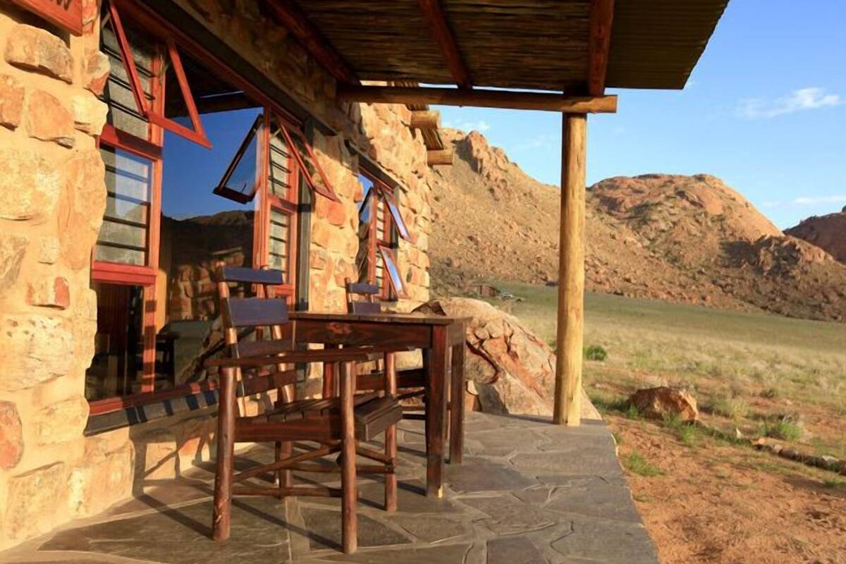 Un edificio de piedra con una mesa y sillas de madera afuera, una montaña al fondo y un cielo despejado.