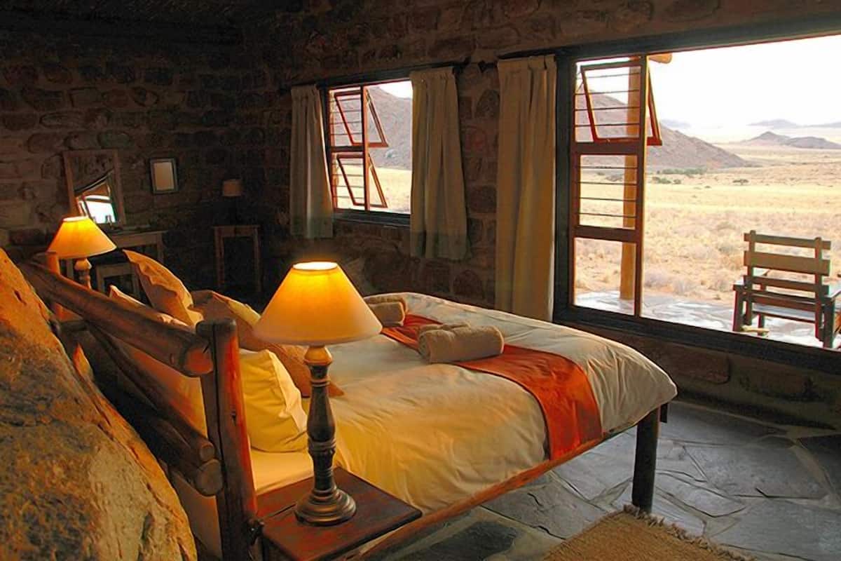 Una habitación con pared de piedra, cama con cabecera de madera, dos lámparas de noche y vista a un paisaje desértico a través de ventanas amplias.