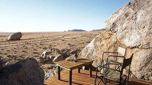 Una terraza de madera con una mesa y sillas, frente a un paisaje desértico con rocas grandes y cielo despejado.