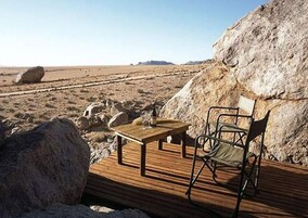 A wooden deck with a table and chairs, set against a desert landscape with large rocks and a clear sky.