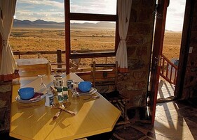 A rustic dining area with a table set for two, a view of a vast landscape through a window, and stone walls.