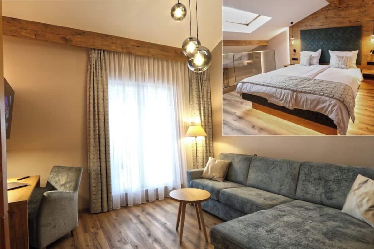 Une chambre d’hôtel avec un plancher en bois, un canapé gris, une petite table ronde, un lit avec une tête de lit grise et une grande fenêtre avec des rideaux.