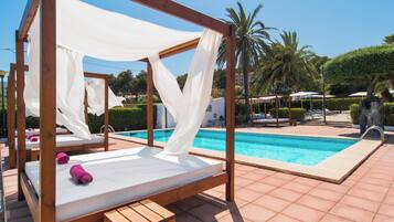 A wooden bed with a white canopy and pink towels, a pool, and palm trees in the background.