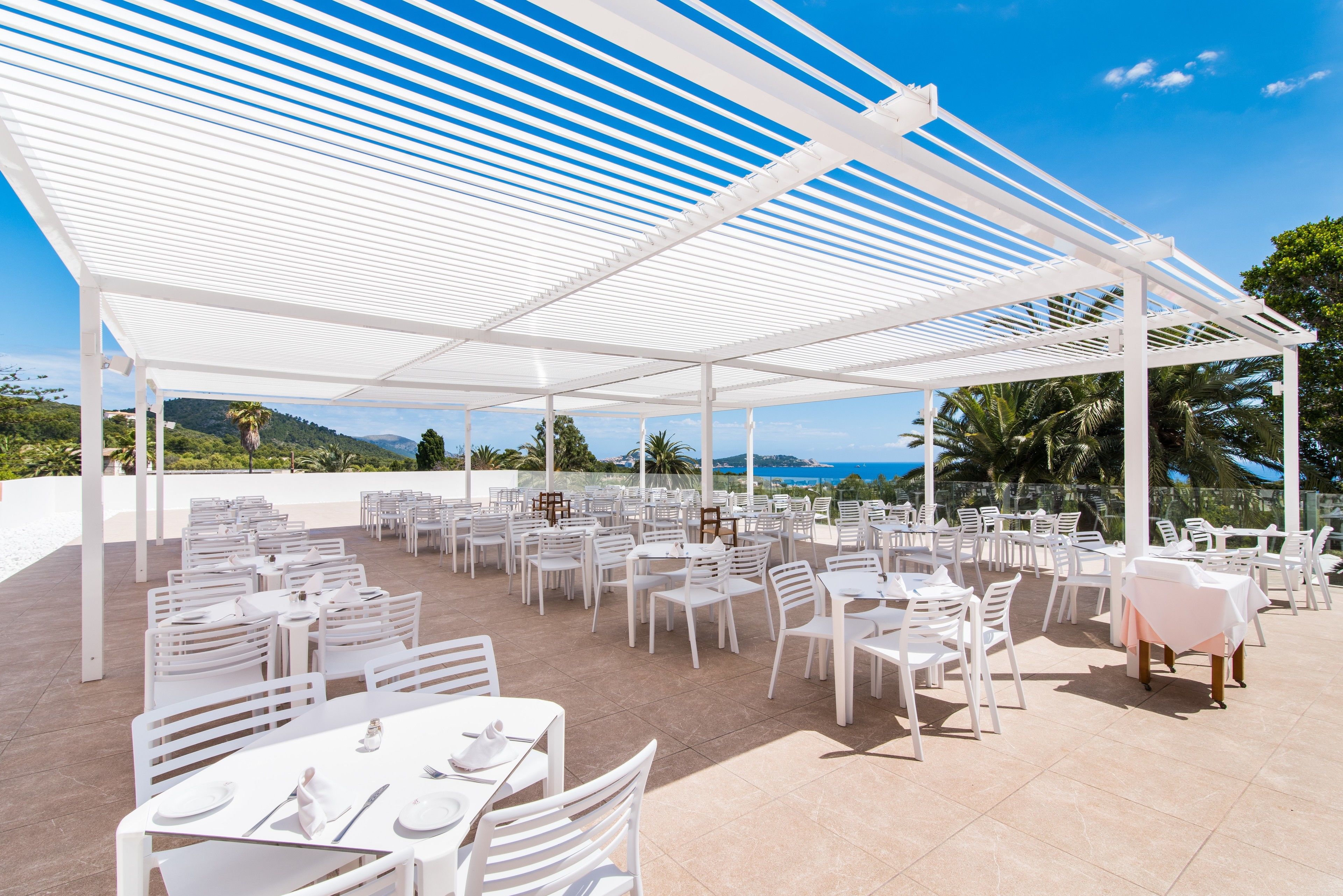 An outdoor dining area with white tables and chairs, covered by a white pergola, overlooking a scenic landscape.