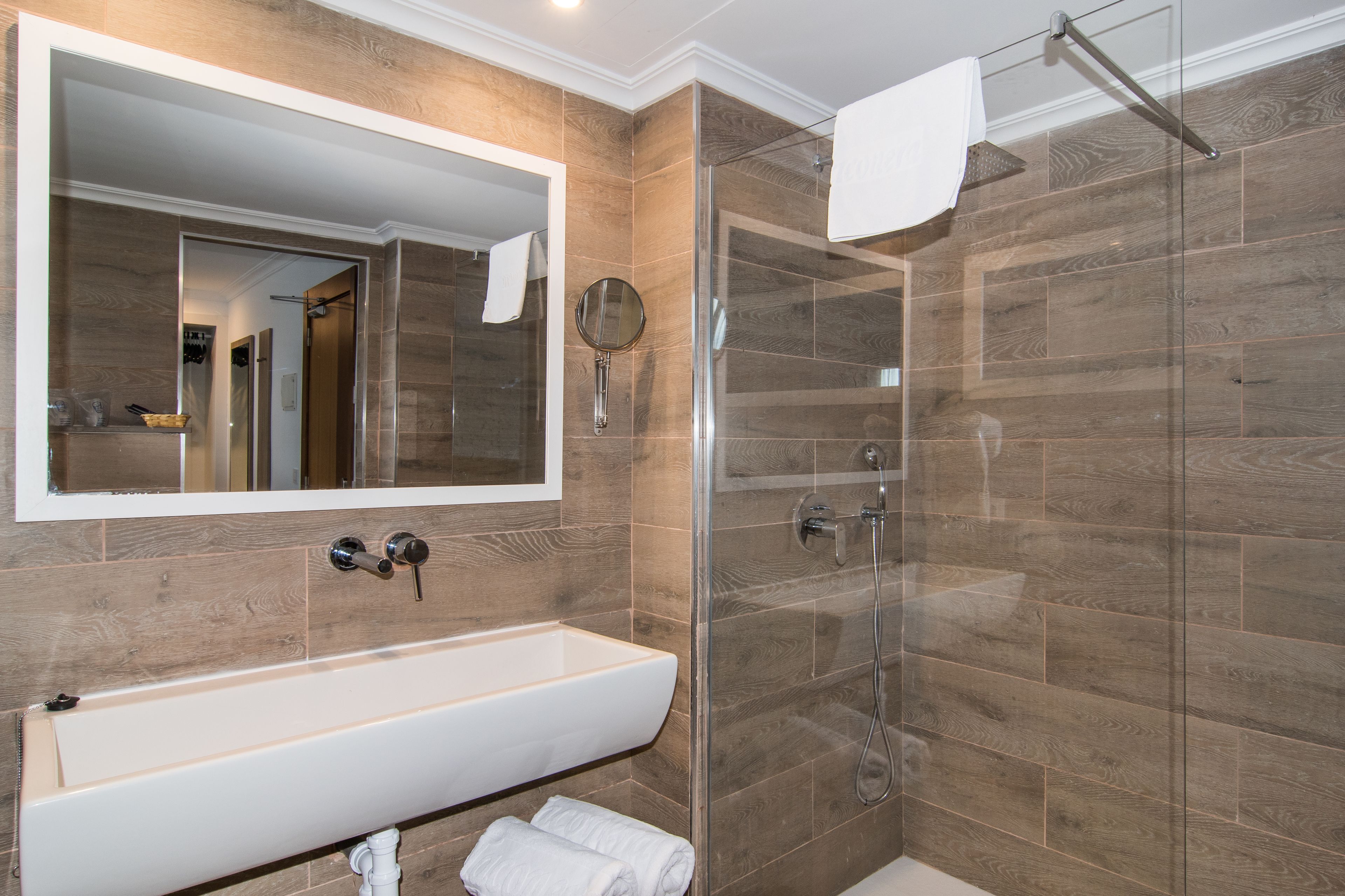 Un baño moderno con un espejo grande, ducha con mampara de vidrio y lavamanos blanco.