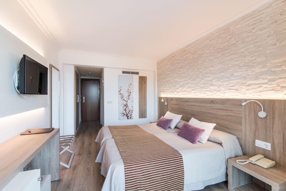 Quarto de hotel moderno com piso de madeira, cama com manta listrada, mesa de cabeceira com luminária e TV de tela plana fixada na parede.