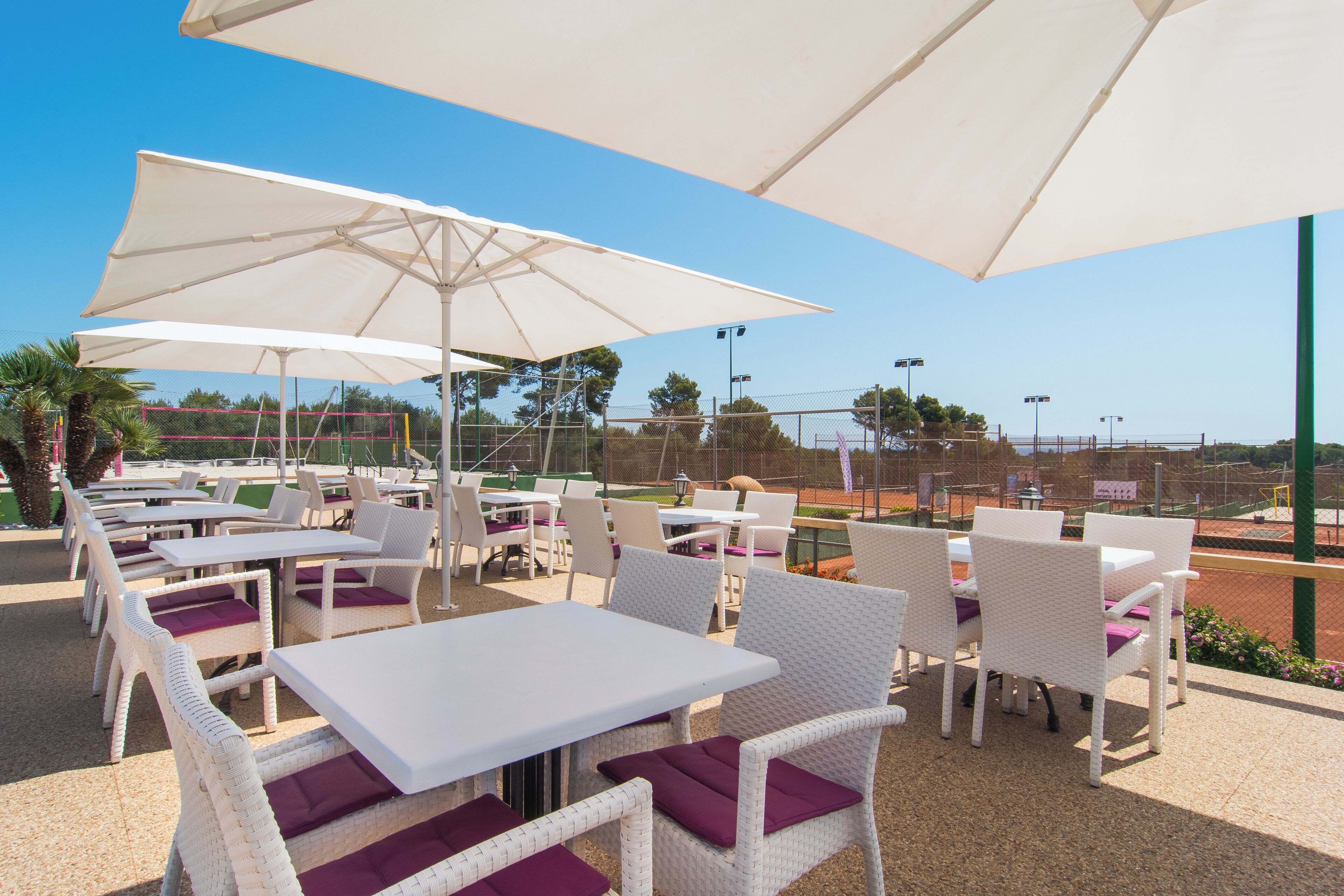An outdoor seating area with white tables and wicker chairs, large umbrellas, and a tennis court in the background.