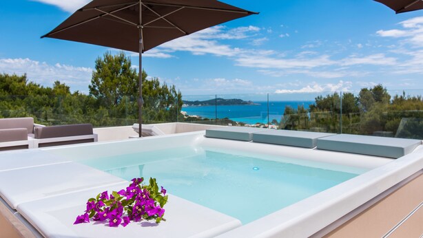 Una piscina infinita en la azotea con sillones de descanso blancos y flores moradas, rodeada de sombrillas grandes y una vista panorámica del océano.
