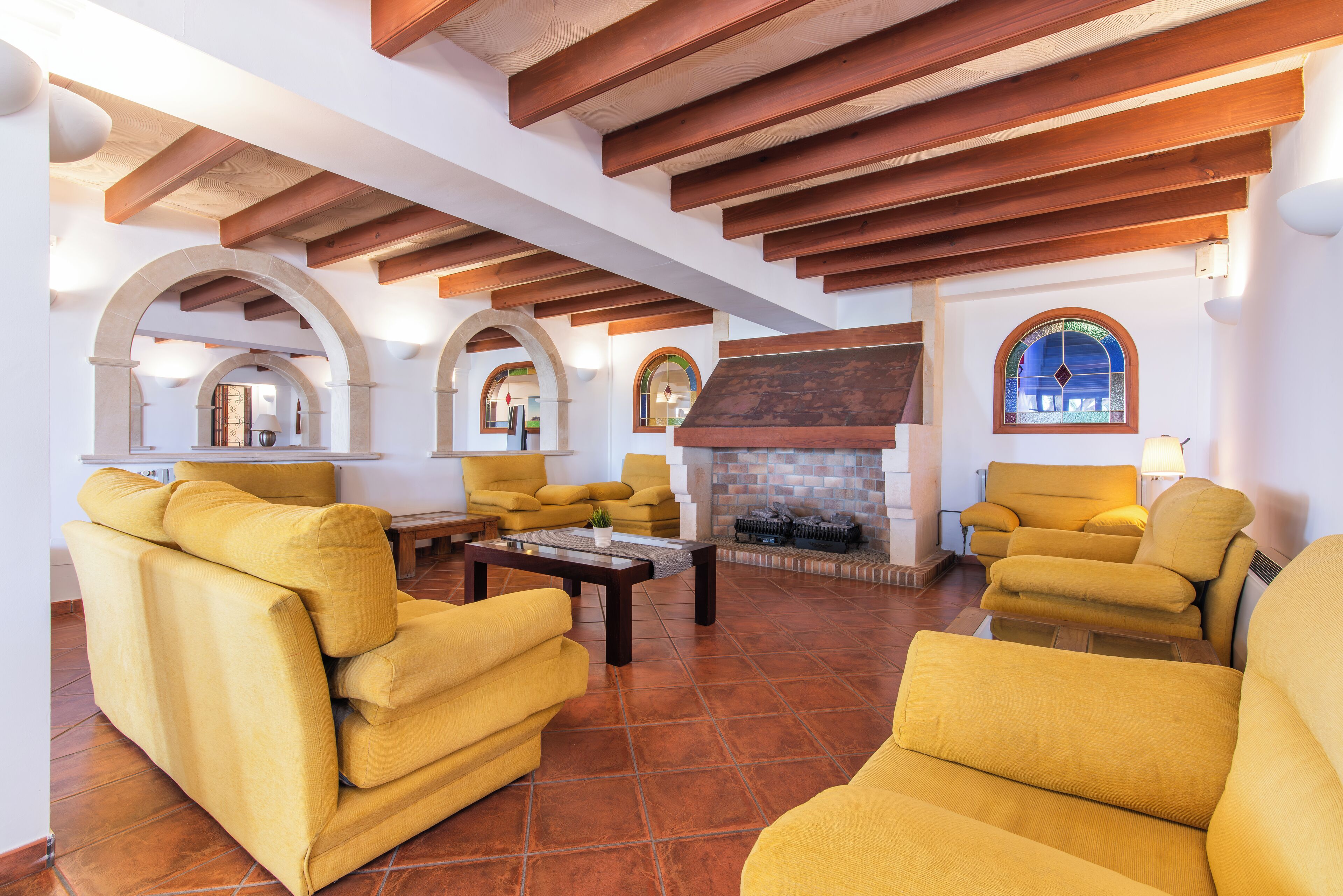 A spacious living room with yellow sofas, a fireplace, and wooden beams on the ceiling.