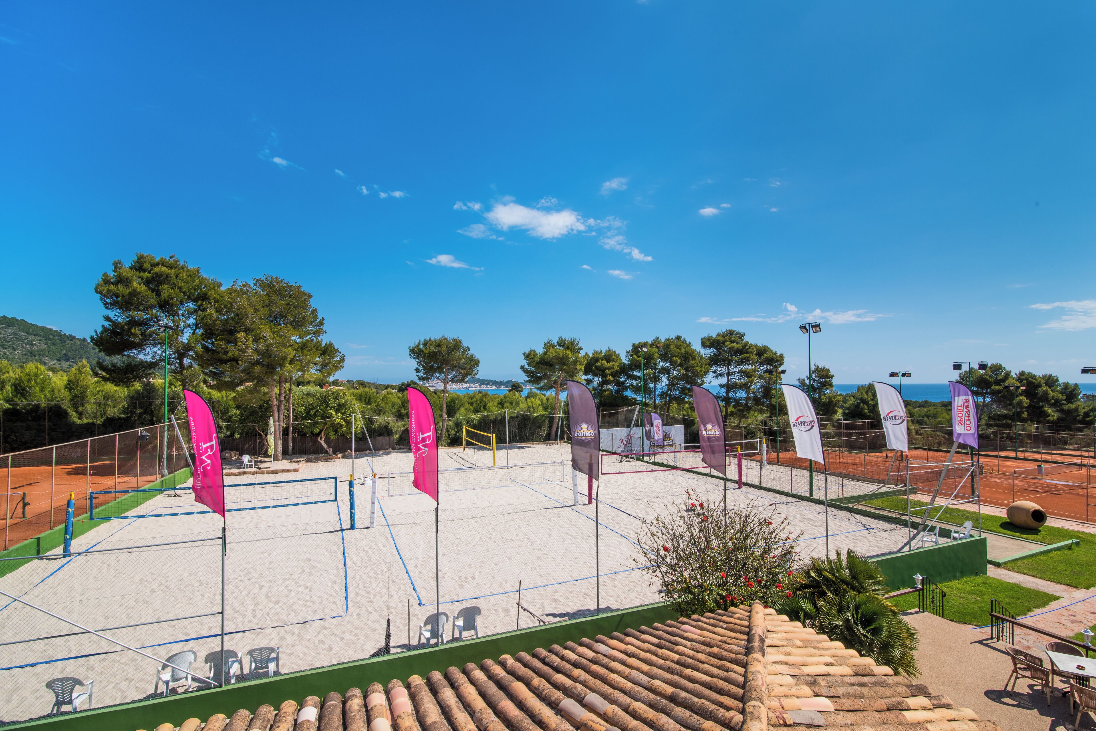 Cancha de tenis con banderas, sillas y un edificio con techo verde.