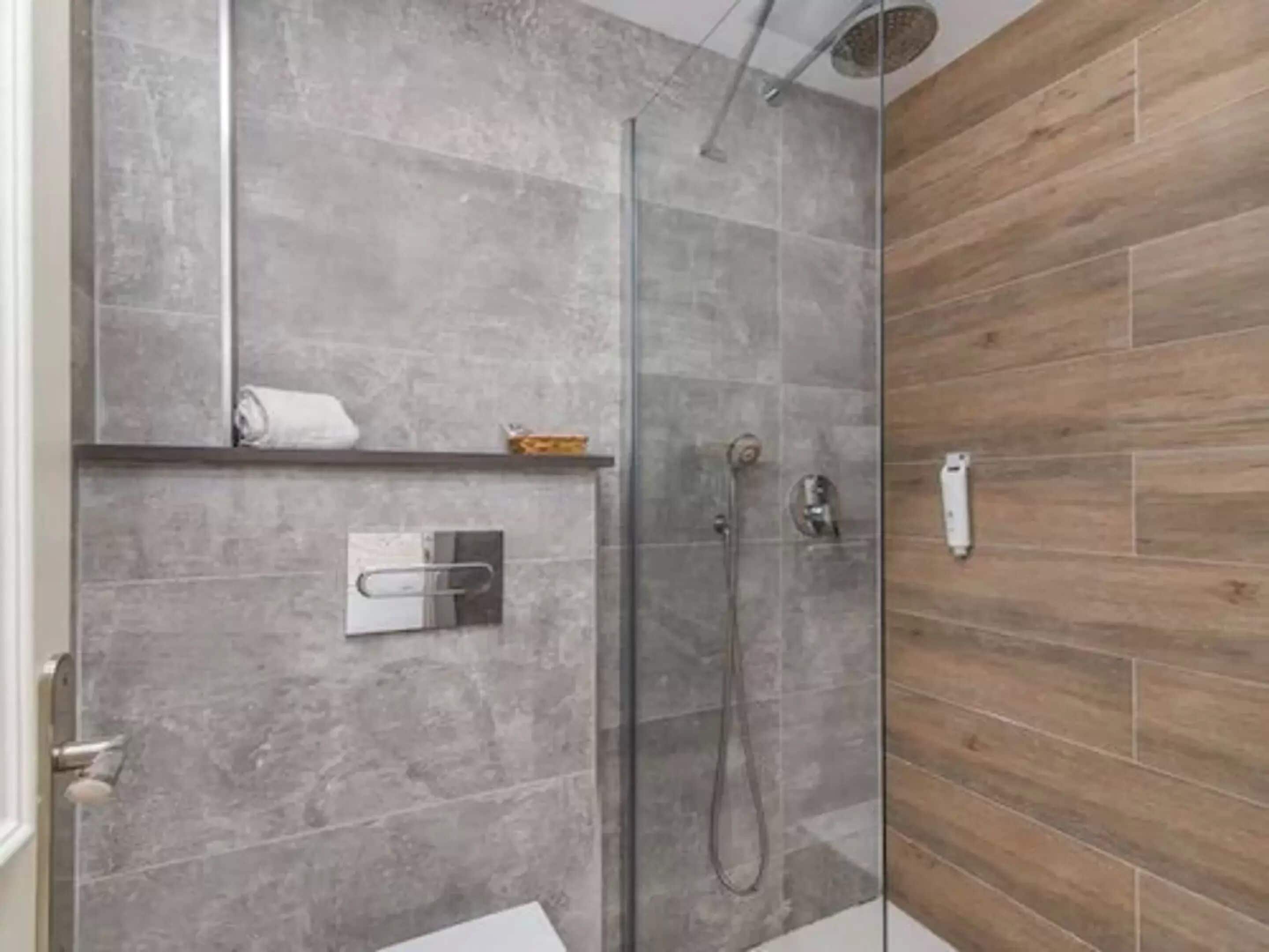 A modern bathroom with a glass shower enclosure, grey tiled walls, and a wooden accent wall.