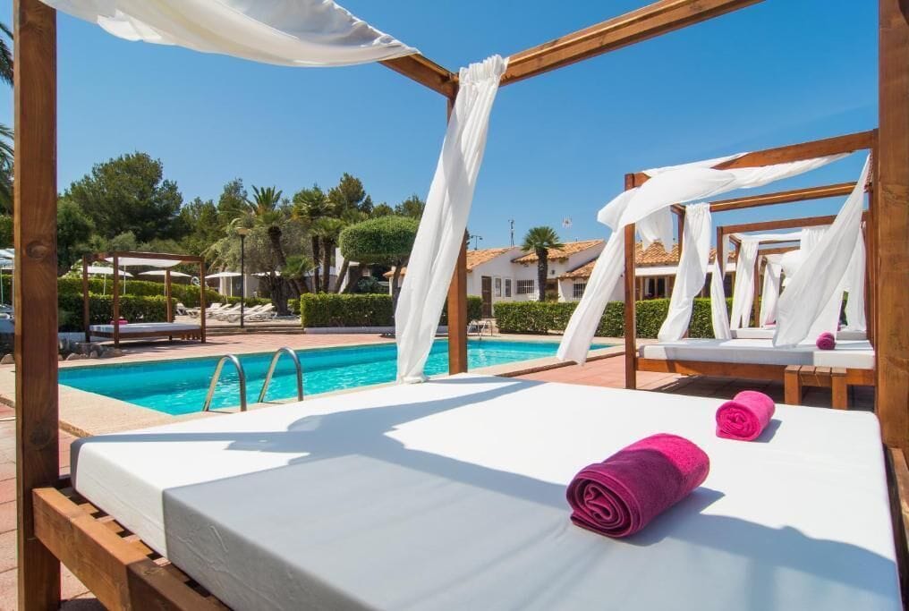 Un letto di legno con biancheria da letto bianca e asciugamani rosa, una piscina e tende bianche in un contesto da resort.