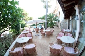 An outdoor patio with wicker furniture and umbrellas, surrounded by greenery and a pool. - Cala Gat (Capdepera)