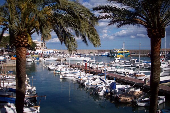 A marina with numerous boats docked, palm trees, and a building with arches in the background. - Baviera Hotel (Capdepera)