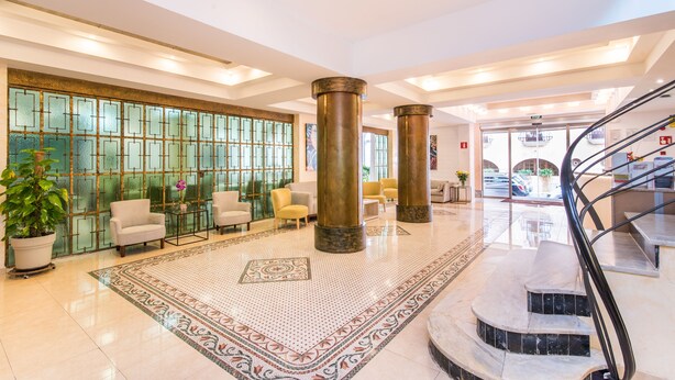 A spacious lobby with a large tiled floor, a seating area with chairs, a staircase, and a decorative wall with glass panels. - Baviera Hotel (Capdepera)