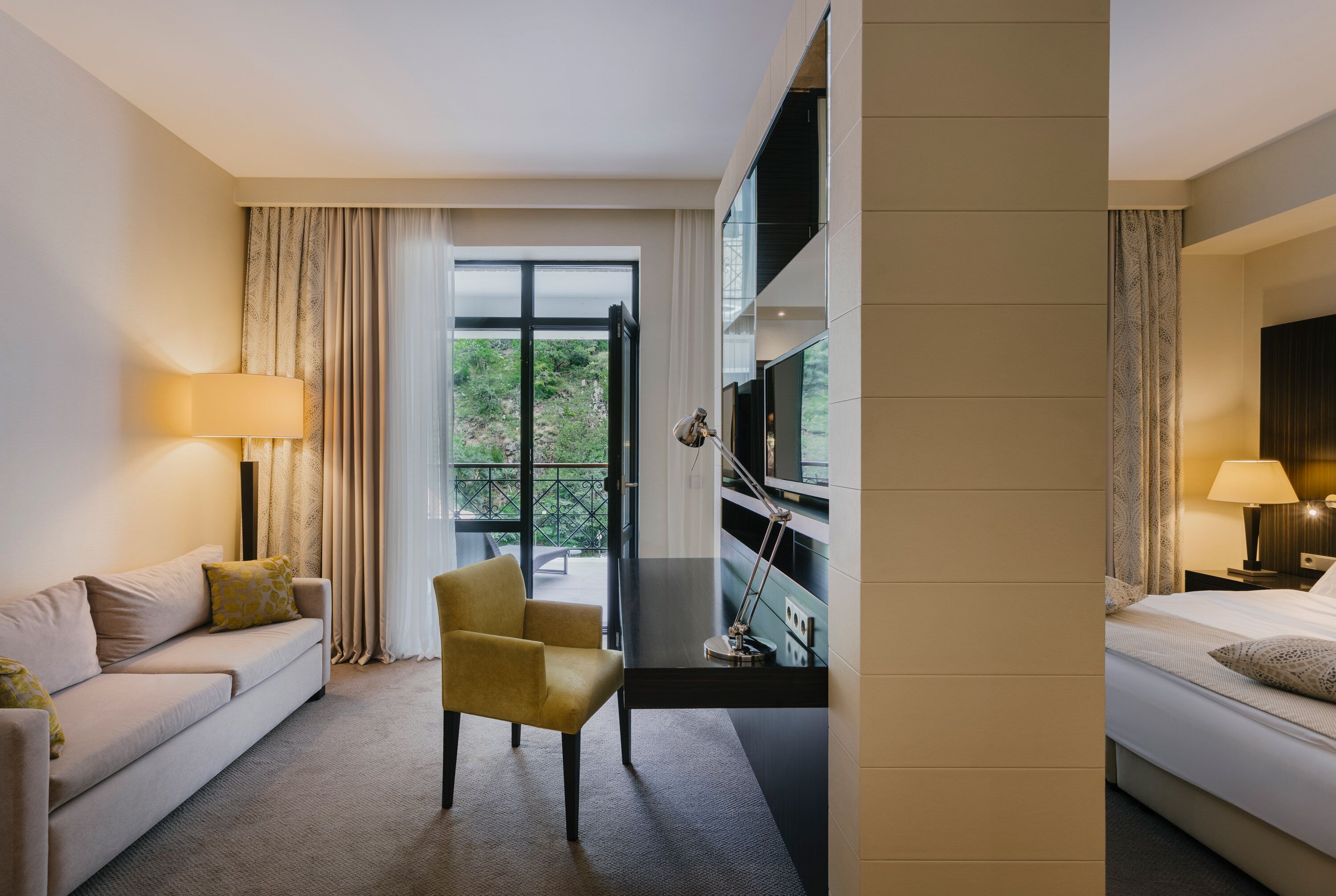 Quarto de hotel com sofá, poltrona, cama, televisão e vista para áreas verdes.