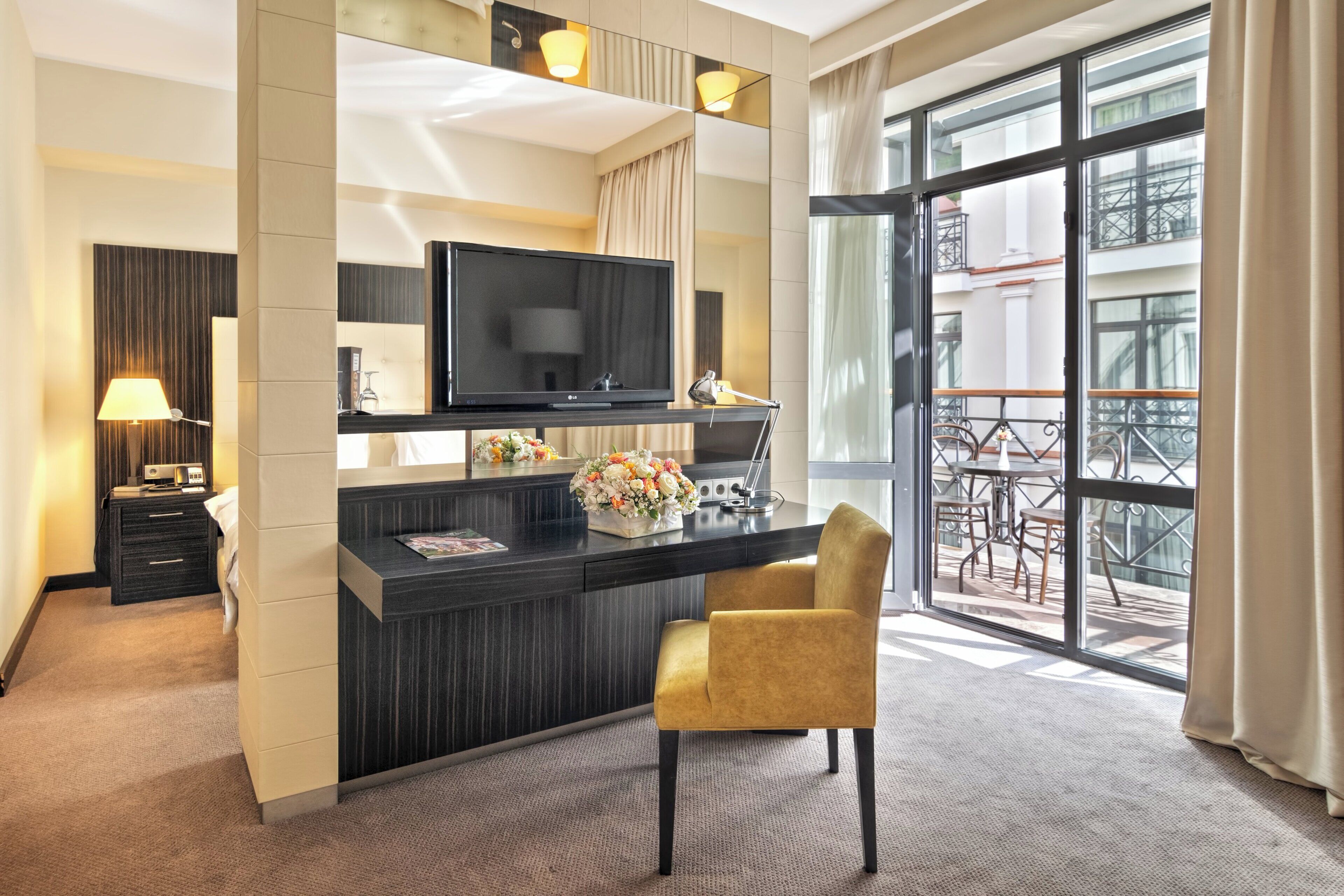 Habitación de hotel con televisión de pantalla plana, escritorio con un jarrón de flores, una silla y balcón con mobiliario exterior.
