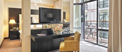 Habitación de hotel con televisión de pantalla plana, escritorio con un jarrón de flores, una silla y balcón con mobiliario exterior.