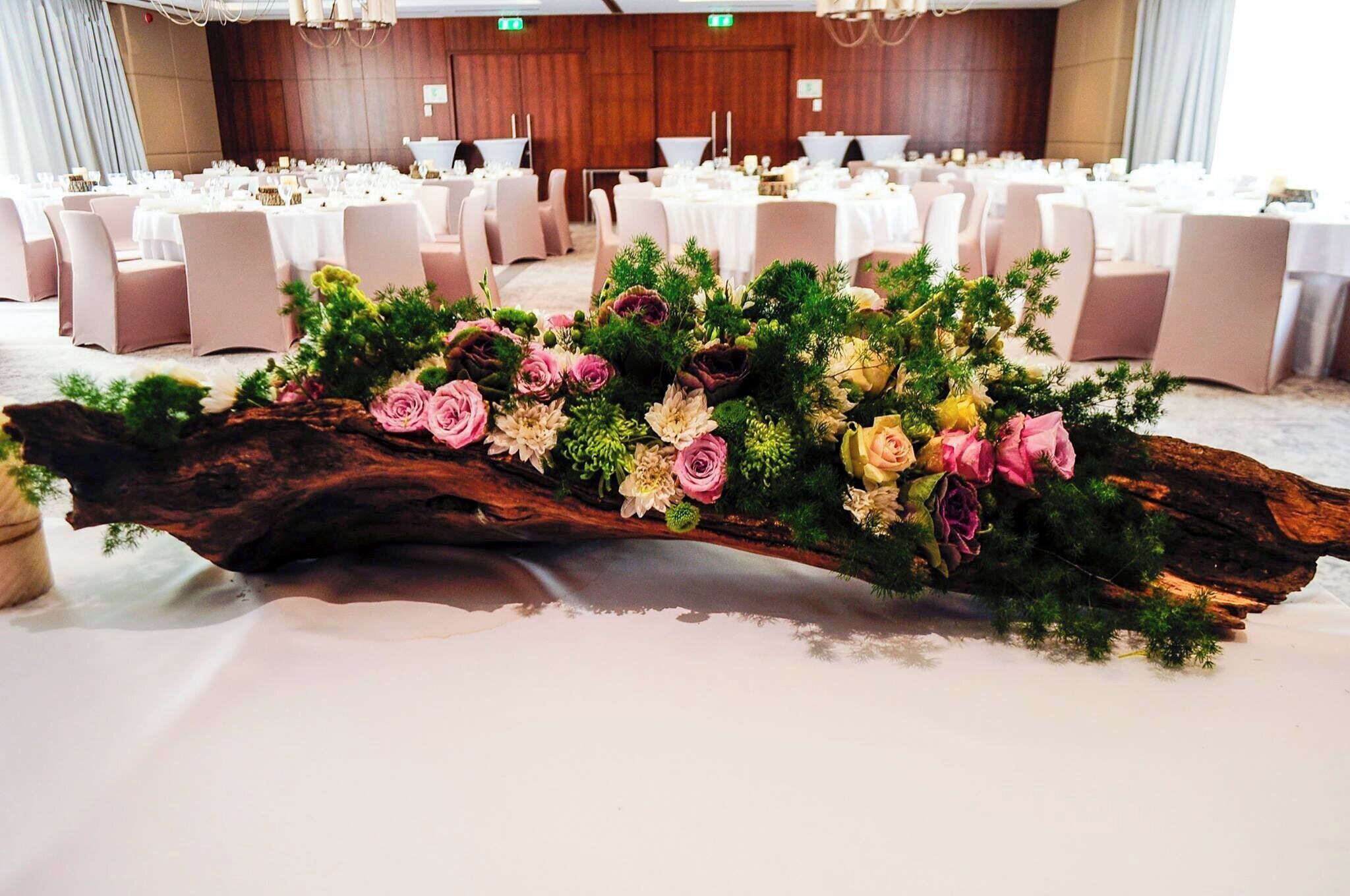 A banquet hall with round tables set for dining, a decorative floral arrangement on a wooden log centerpiece, and a stage with a podium in the background.