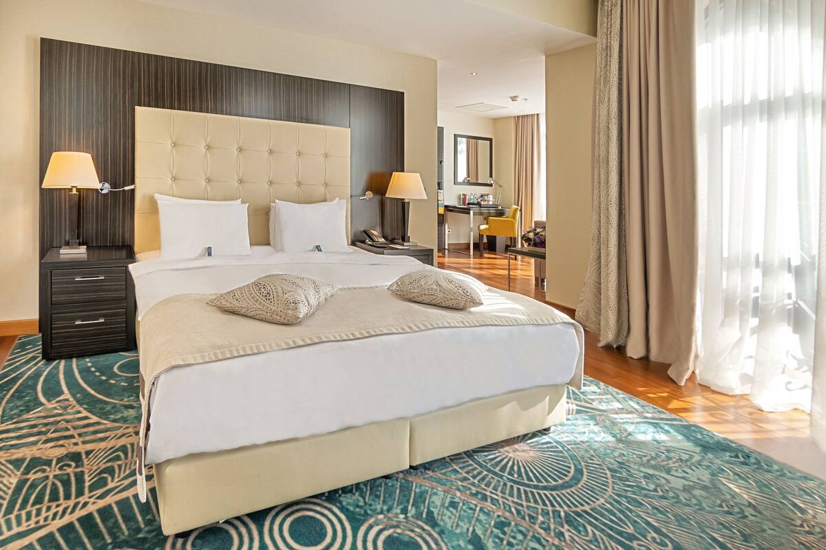 Habitación de hotel con una cama grande, dos mesitas de noche con lámparas, un escritorio con silla, un espejo y vistas al exterior a través de cortinas translúcidas.