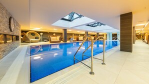 Piscina interna com claraboia, cadeiras de descanso e parede de pedra.