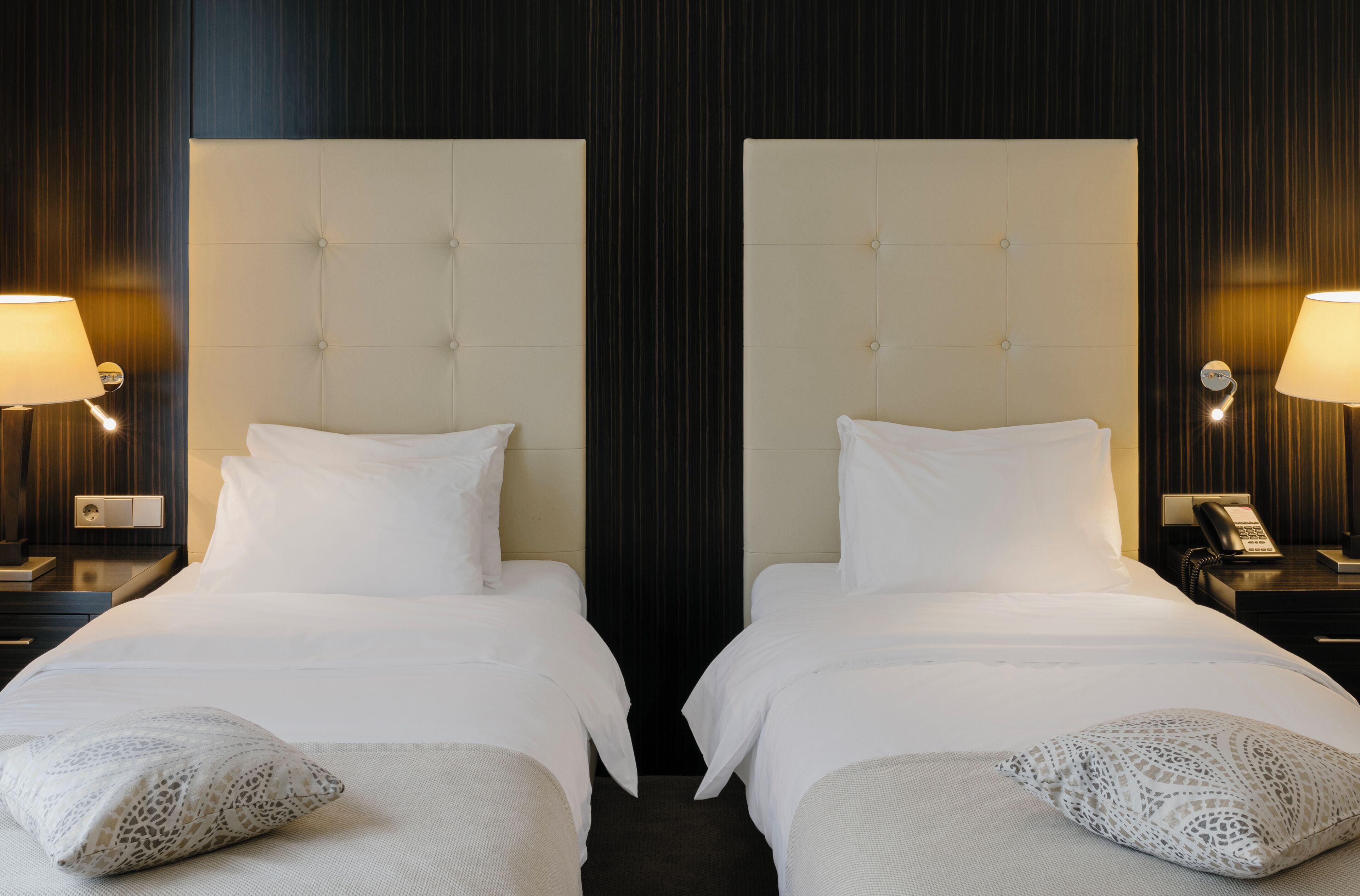 Habitación de hotel con dos camas, cada una con cabecero blanco y almohadas a juego. Se observan mesitas de noche con lámparas y un teléfono a un lado.