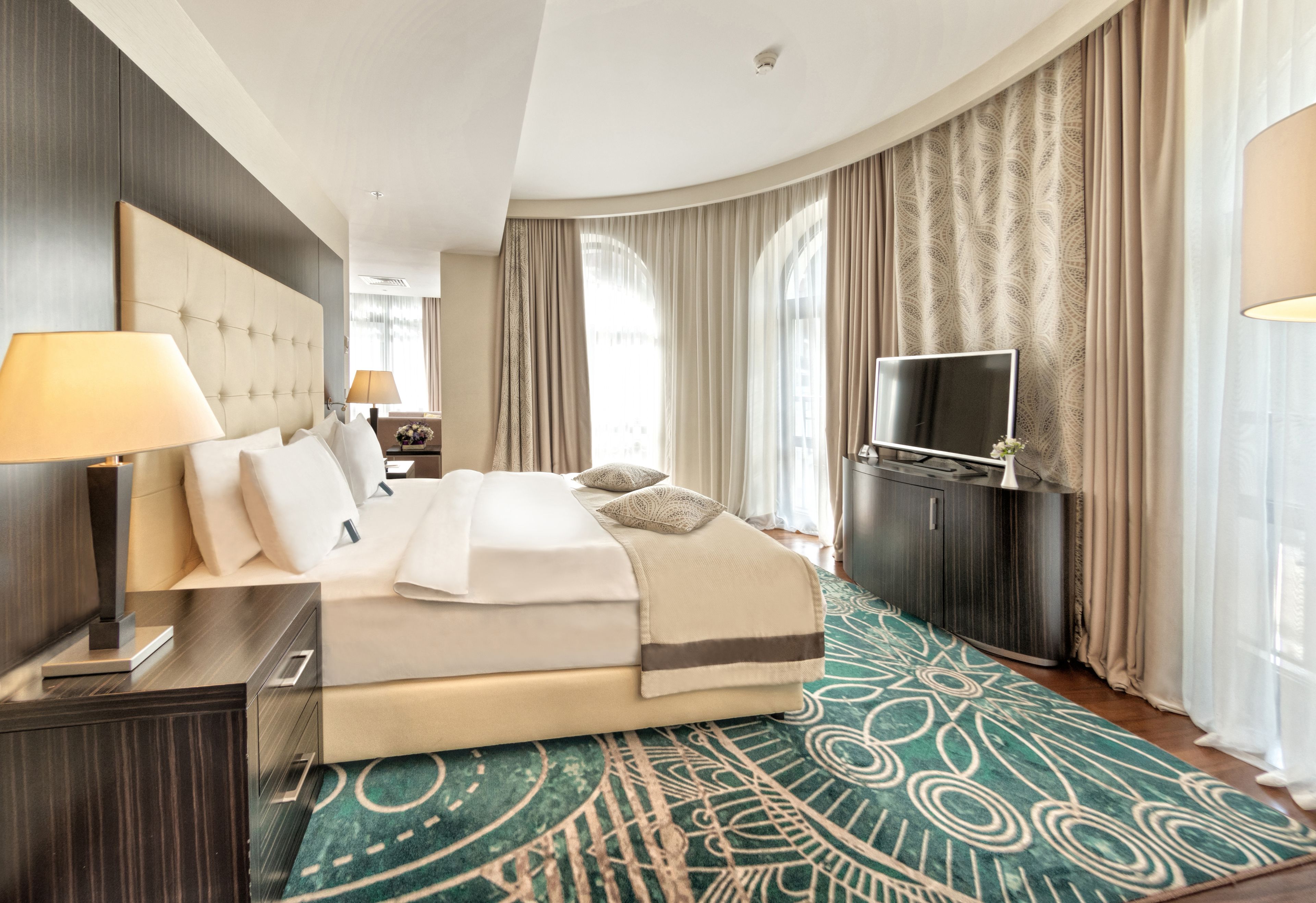 Una habitación de hotel con una cama grande, un televisor, un escritorio con lámpara y una alfombra estampada.