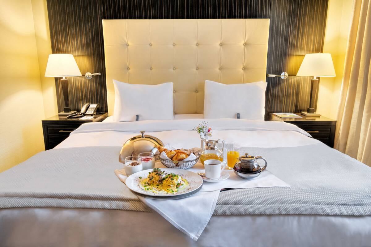 Habitación de hotel con una cama grande, dos mesitas de noche, lámparas de noche, un teléfono y una bandeja de desayuno con cruasanes, café y zumo de naranja.