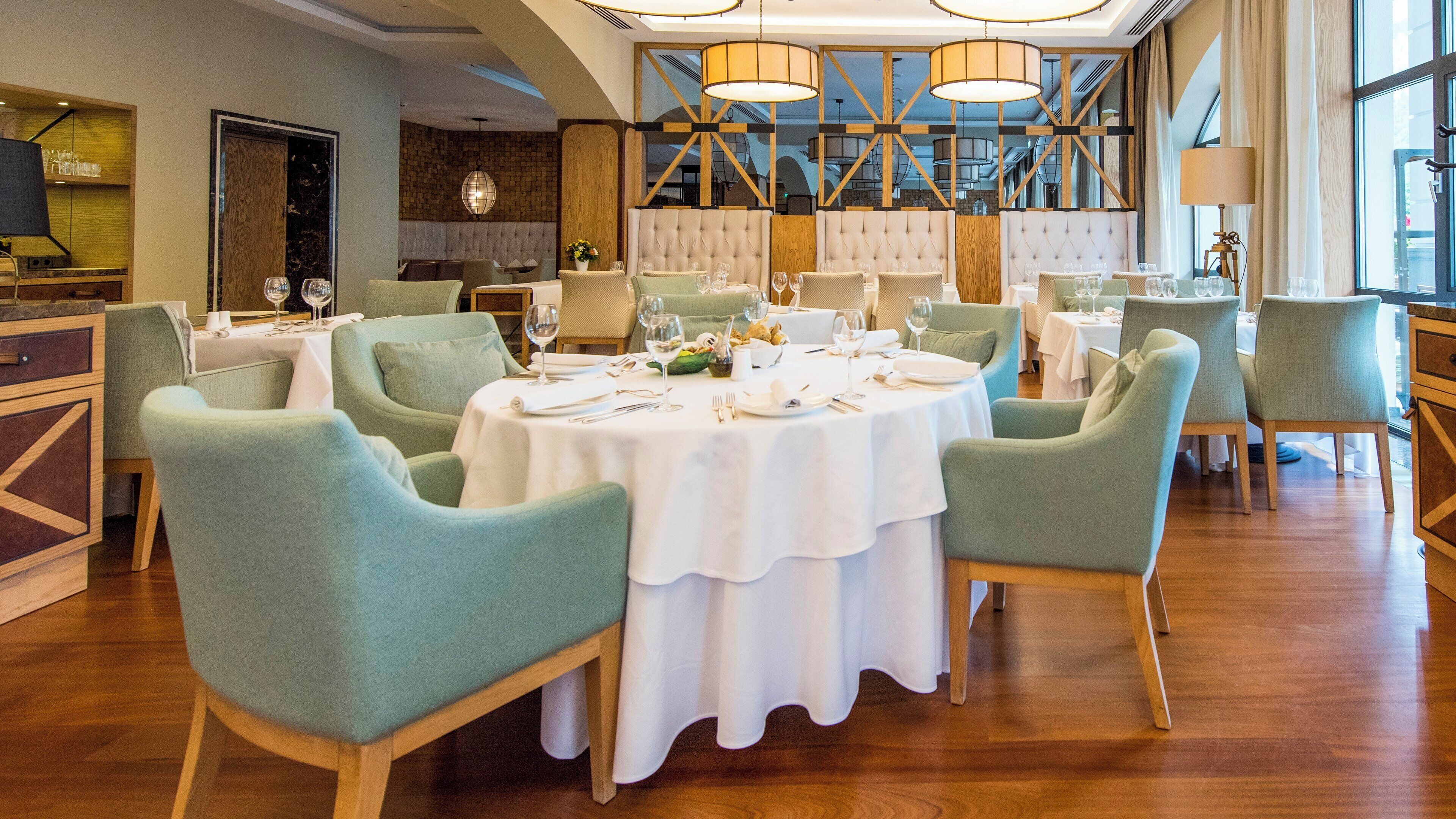 A restaurant with round tables, green upholstered chairs, and a wooden floor.