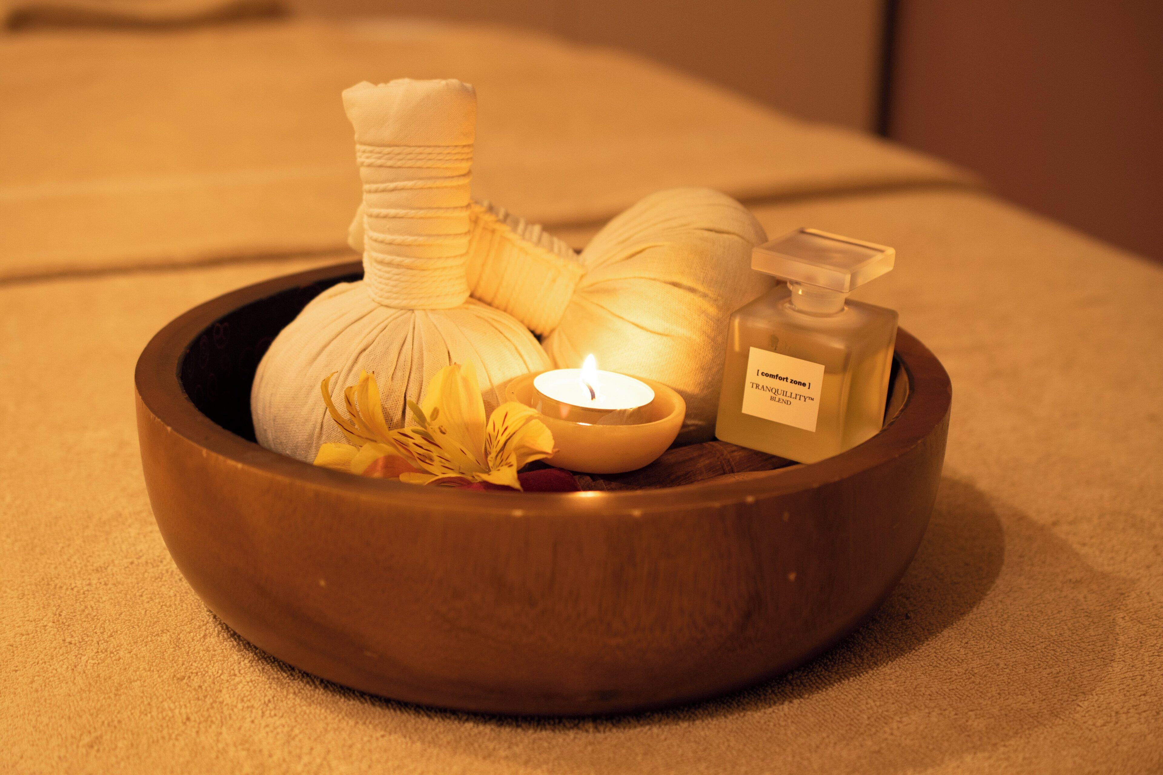 A wooden bowl containing a massage stone, a candle, and a bottle of essential oil.