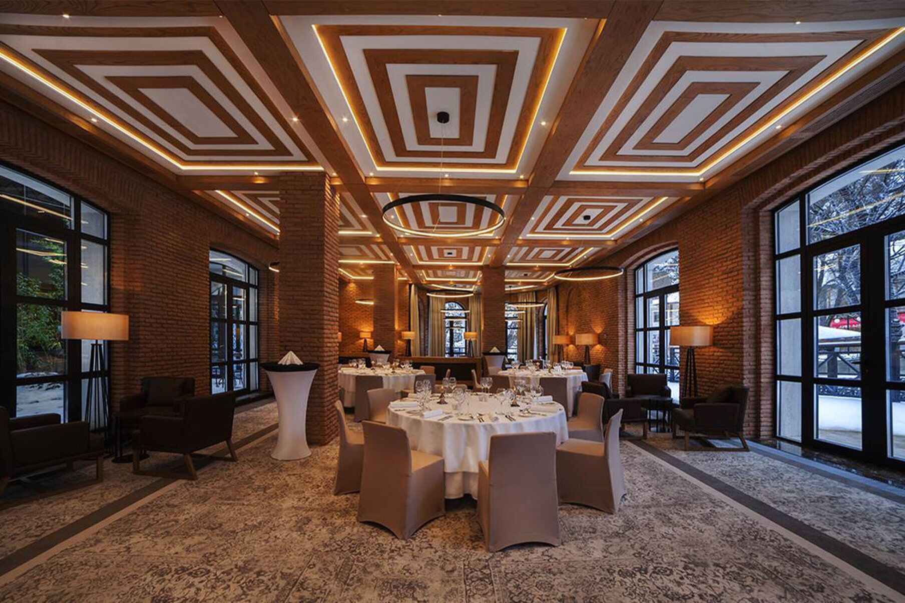 A spacious banquet hall with a high ceiling featuring geometric patterns, large windows, and round tables set for dining.