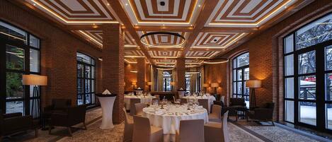 A spacious banquet hall with a high ceiling featuring geometric patterns, large windows, and round tables set for dining.