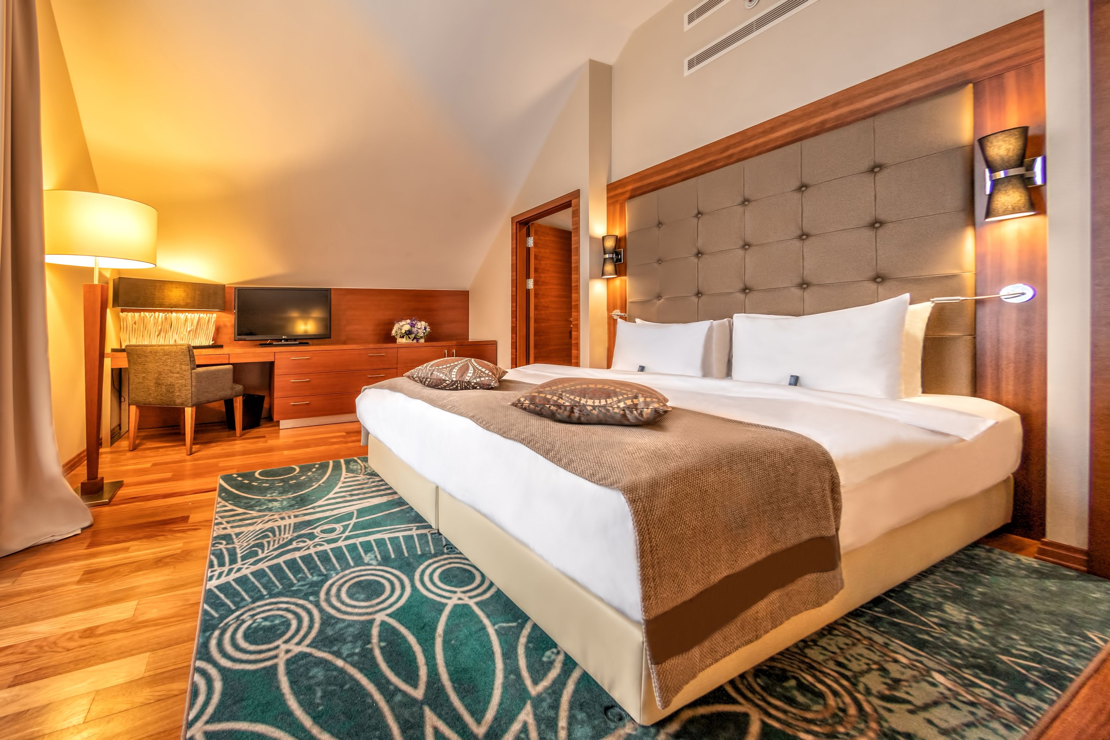 Habitación de hotel con una cama grande, un escritorio con televisor, una silla y un cabecero de madera.
