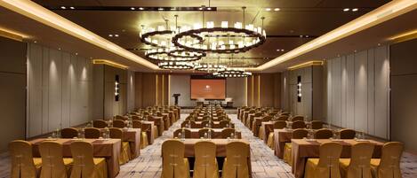 A conference room with rows of tables and chairs, a large screen at the front, and a modern chandelier.