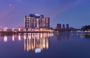 A modern hotel with a distinctive facade, situated by a calm body of water reflecting its lights.