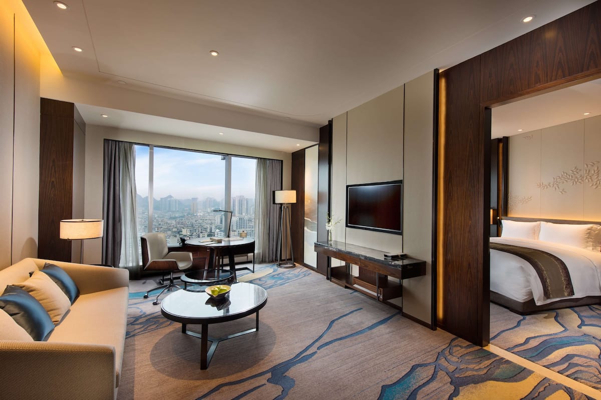 Habitación de hotel con una cama grande, televisión, zona de estar con mesa y sillas, y vistas a la ciudad a través de amplios ventanales.