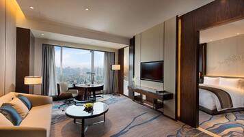 Ein Hotelzimmer mit einem großen Bett, einem Fernseher, einer Sitzecke mit Tisch und Stühlen sowie Blick auf die Stadt durch große Fenster.