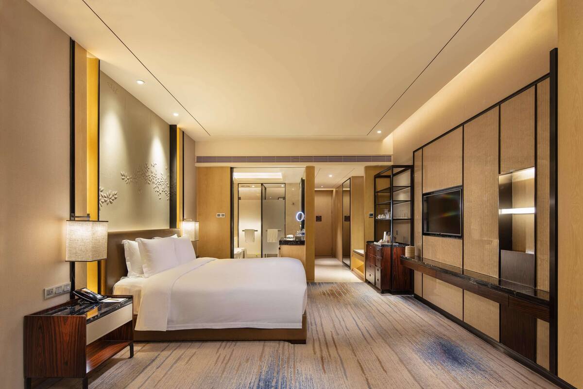 Habitación de hotel con una cama grande, mesitas de noche, televisión, espejo y un baño visible a través de una puerta abierta.