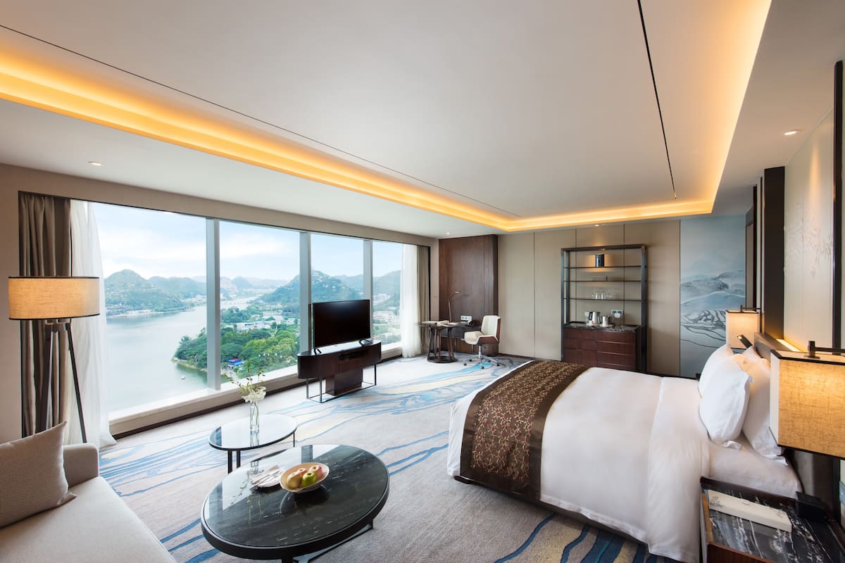 Una habitación de hotel moderna con una cama grande, un televisor de pantalla plana, un escritorio con una silla y vistas a un río y montañas.